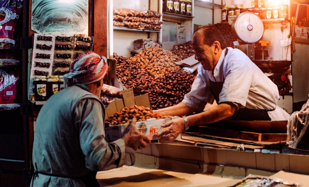 man-in-blue-top-giving-box-to-man-in-gray-top-696205 Two men exchanging dates in a vibrant Dubai spice market stall.